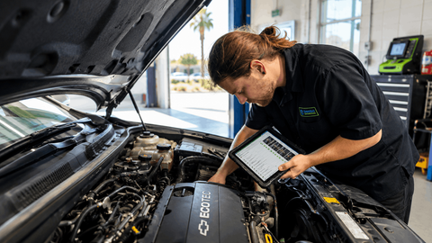 Professional automotive technician performing a digital vehicle inspection under the hood of a modern vehicle inside a clean Phoenix auto repair shop, using a tablet inspection checklist with Arizona sunlight visible in the background.
